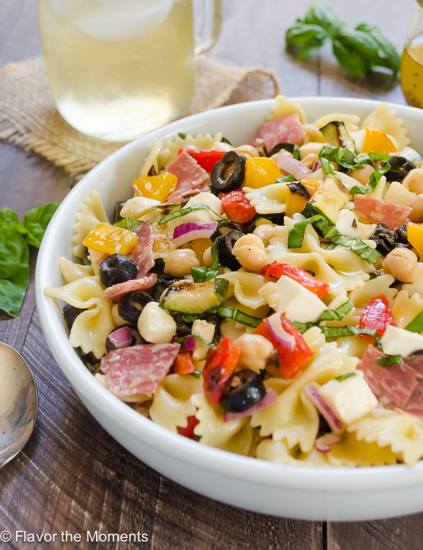 A white serving bowl filled with zesty Italian pasta salad. A glass of tea rests in the background. 