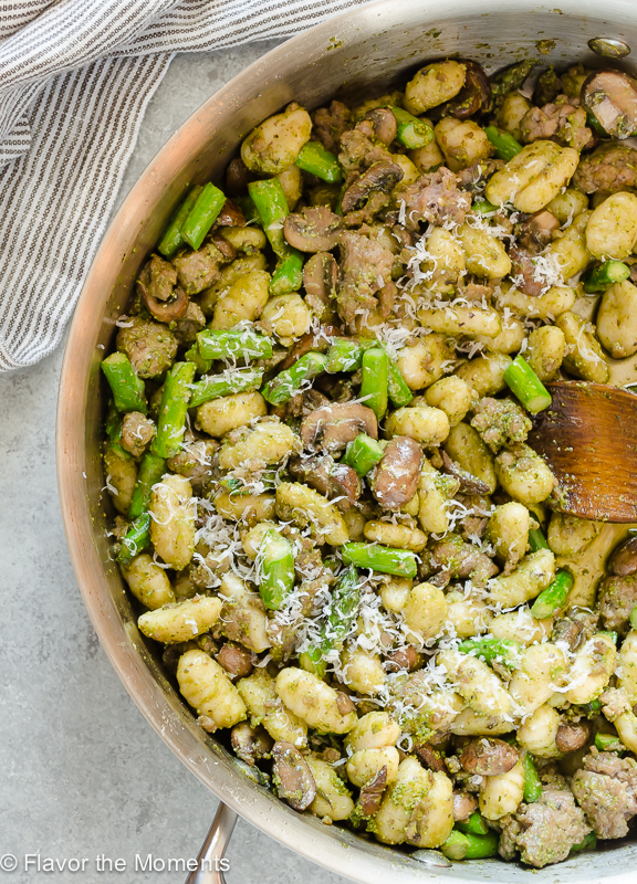 Overhead view of pesto gnocchi with sausage and veggies in a skillet. A wood spatula rests in the skillet. 