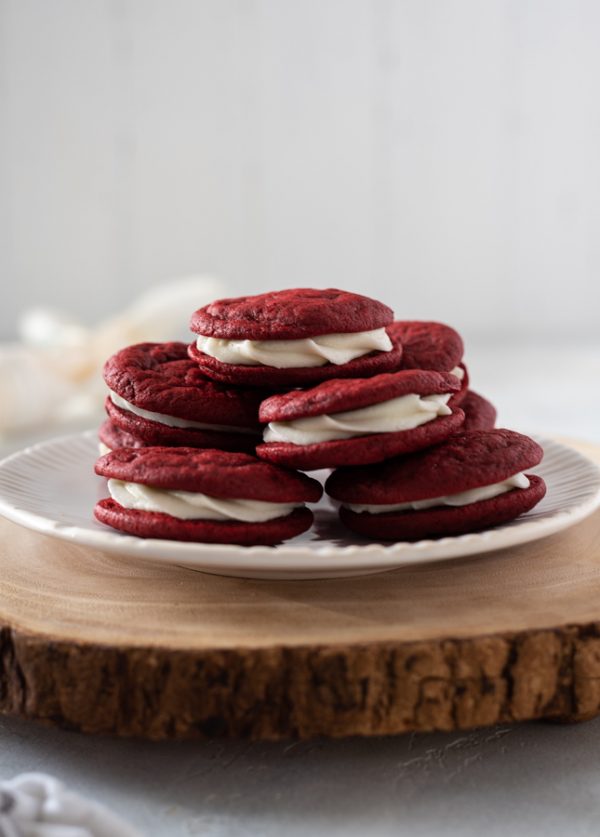 Red velvet sandwich cookies piled high on white plate atop a wooden server