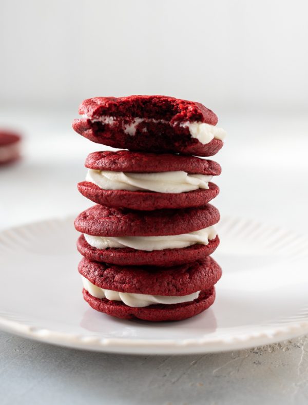 Red velvet sandwich cookies stacked up on white plate with bite out of top cookie