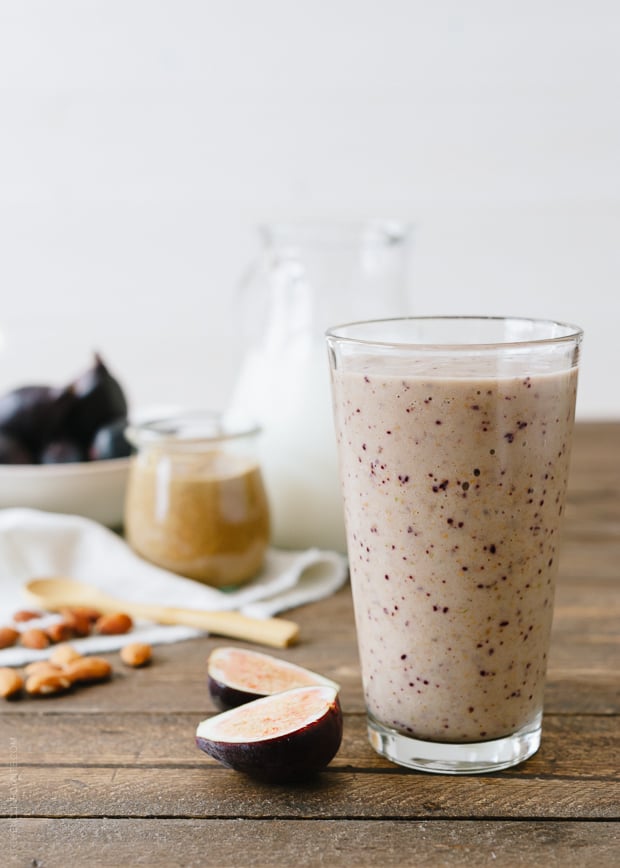 A fig and almond butter smoothie in a tall glass. Figs, almond butter, and almonds rest in the background.