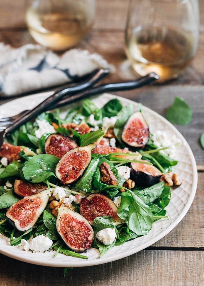 A fresh fig salad on a large white plate. Two glasses of wine rest in the background.