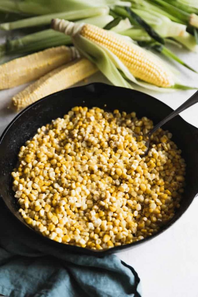 A cast iron skillet of fried corn, next to fresh ears of corn.