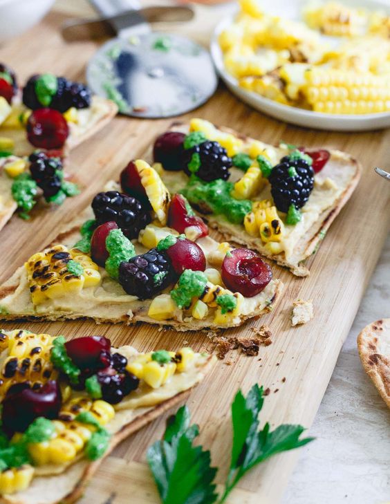 Sliced hummus flatbread with corn and berries on a wood serving board.