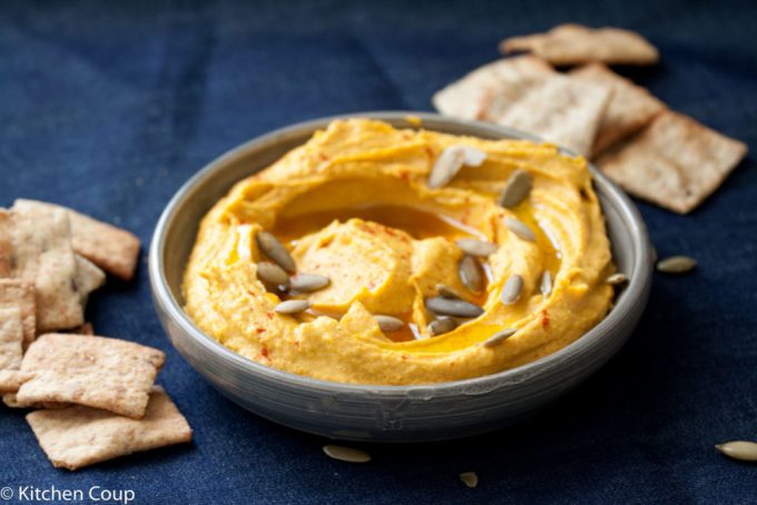 A dish of kabocha squash hummus surrounded by pita chips, on a dark blue background.