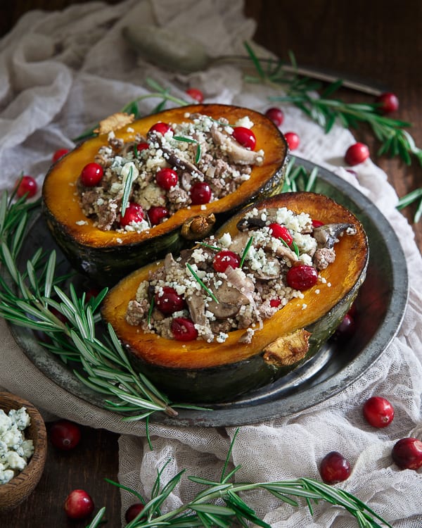 Two lamb-stuffed kabocha squash halves on a metal plate. Fresh rosemary and cranberries surround the plate.