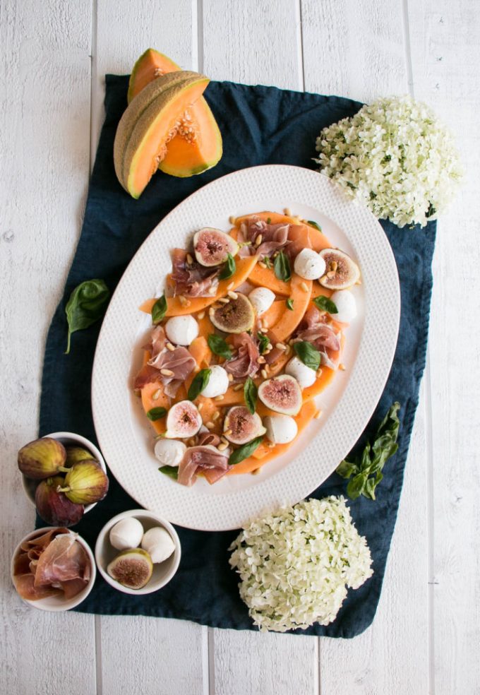 Overhead view of a melon prosciutto salad on a white platter. Individual ingredients surround the platter on a blue towel.
