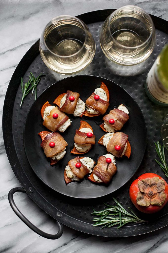 Overhead view of persimmon bacon bites on a gray plate, next to two glasses of wine.