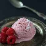 A scoop of rhubarb ice cream with fresh raspberries on a dark plate. A spoon rests in the background.