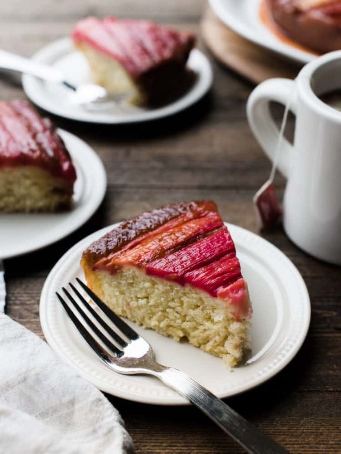Rhubarb upside down cake on a white plate with a fork. Two more slices of cake rest on plates behind it, as does a coffee mug.