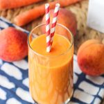 A peach mango carrot smoothie in a glass with two paper striped straws.