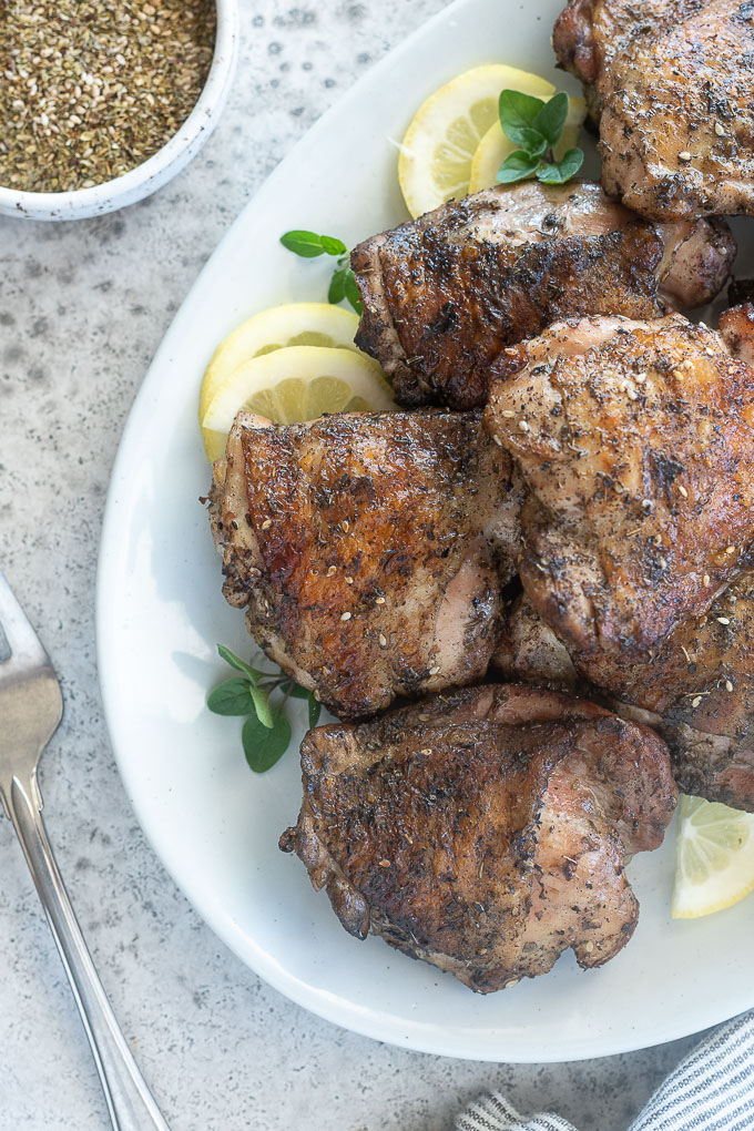 Grilled chicken thighs piled on a platter with lemon and oregano