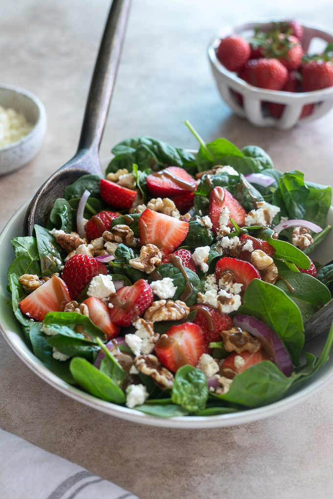Spinach and strawberry salad in a white bowl with serving spoon
