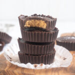 Stack of protein peanut butter cups on a cutting board with flaky sea salt scattered around.