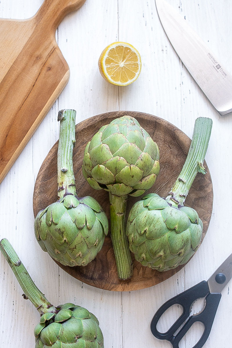 Artichokes with knife lemon and tools for prepping on cutting board.