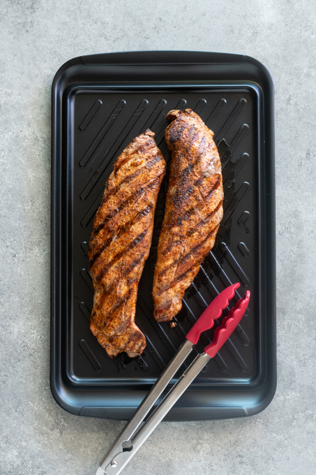 Grilled pork tenderloins resting on cutting board with tongs.
