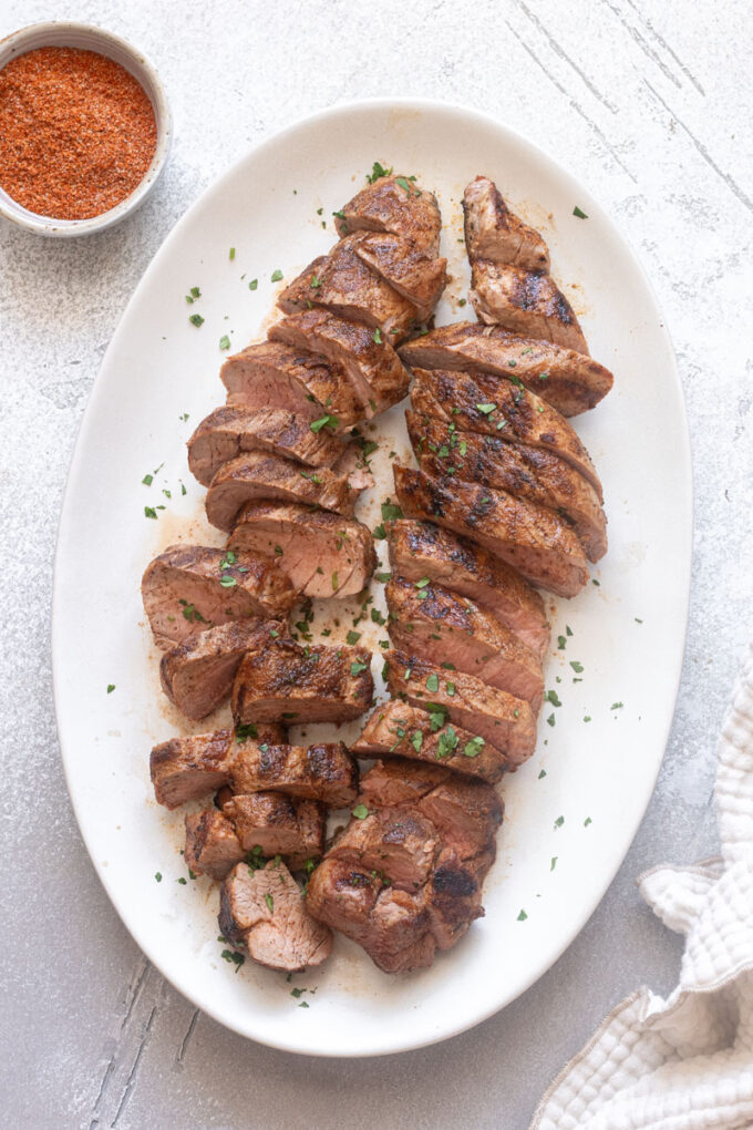 Grilled pork tenderloin sliced on a platter with spice rub in bowl.
