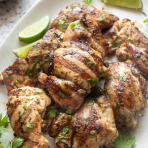 Grilled cilantro lime chicken thighs on a white serving platter garnished with fresh cilantro and lime wedges.