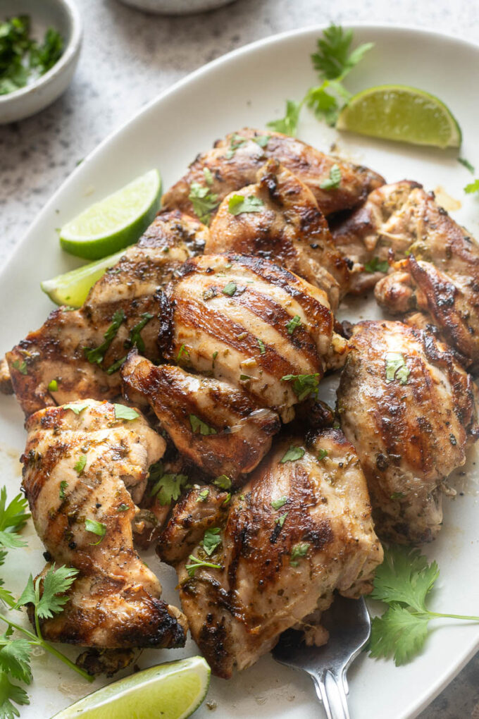 Grilled cilantro lime chicken thighs on a white serving platter garnished with fresh cilantro and lime wedges.