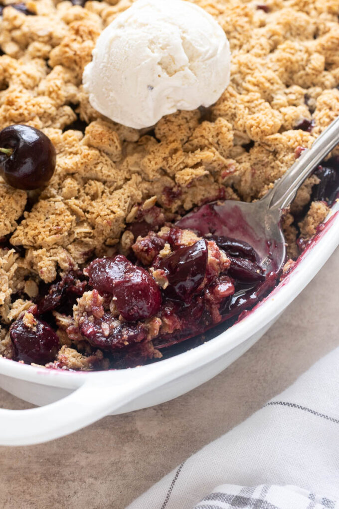 Spoonful of fresh cherry crisp in baking dish with ice cream.