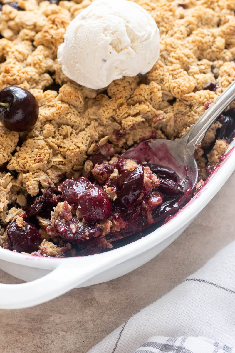 Spoonful of fresh cherry crisp in baking dish with ice cream.