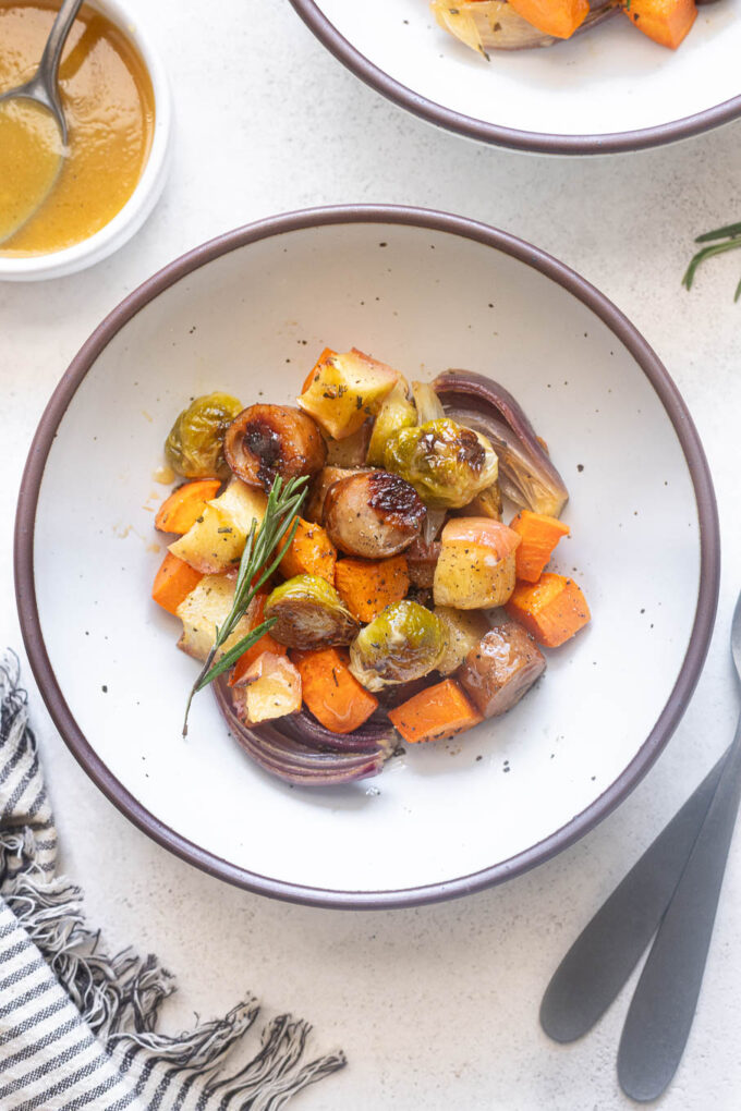Overhead view of a bowl of roasted chicken apple sausage and fall vegetables with rosemary and maple Dijon sauce on the side.