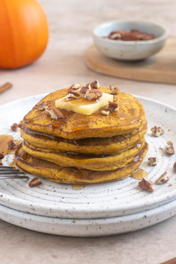 Four golden pumpkin oat flour pancakes stacked with butter, maple syrup and pecans.