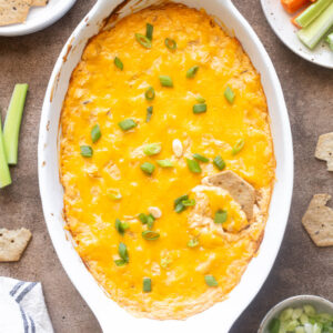 High protein buffalo chicken dip in a white baking dish with crackers and veggies.