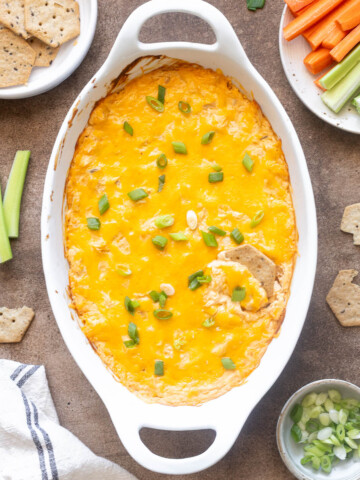 High protein buffalo chicken dip in a white baking dish with crackers and veggies.