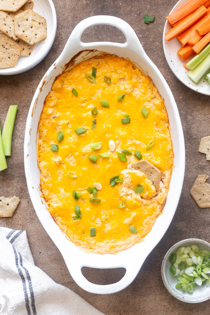 High protein buffalo chicken dip in a white baking dish with crackers and veggies.