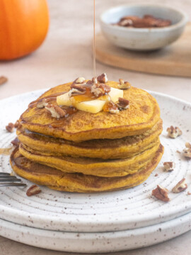 Maple syrup being poured over a stack of fluffy pumpkin oat flour pancakes topped with butter and chopped pecans.