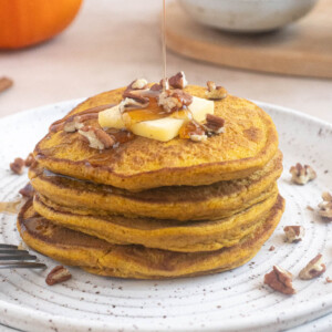 Maple syrup being poured over a stack of fluffy pumpkin oat flour pancakes topped with butter and chopped pecans.