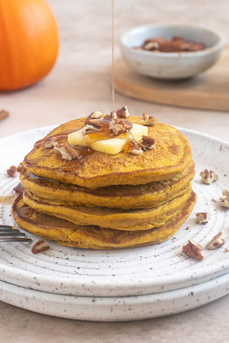Maple syrup being poured over a stack of fluffy pumpkin oat flour pancakes topped with butter and chopped pecans.
